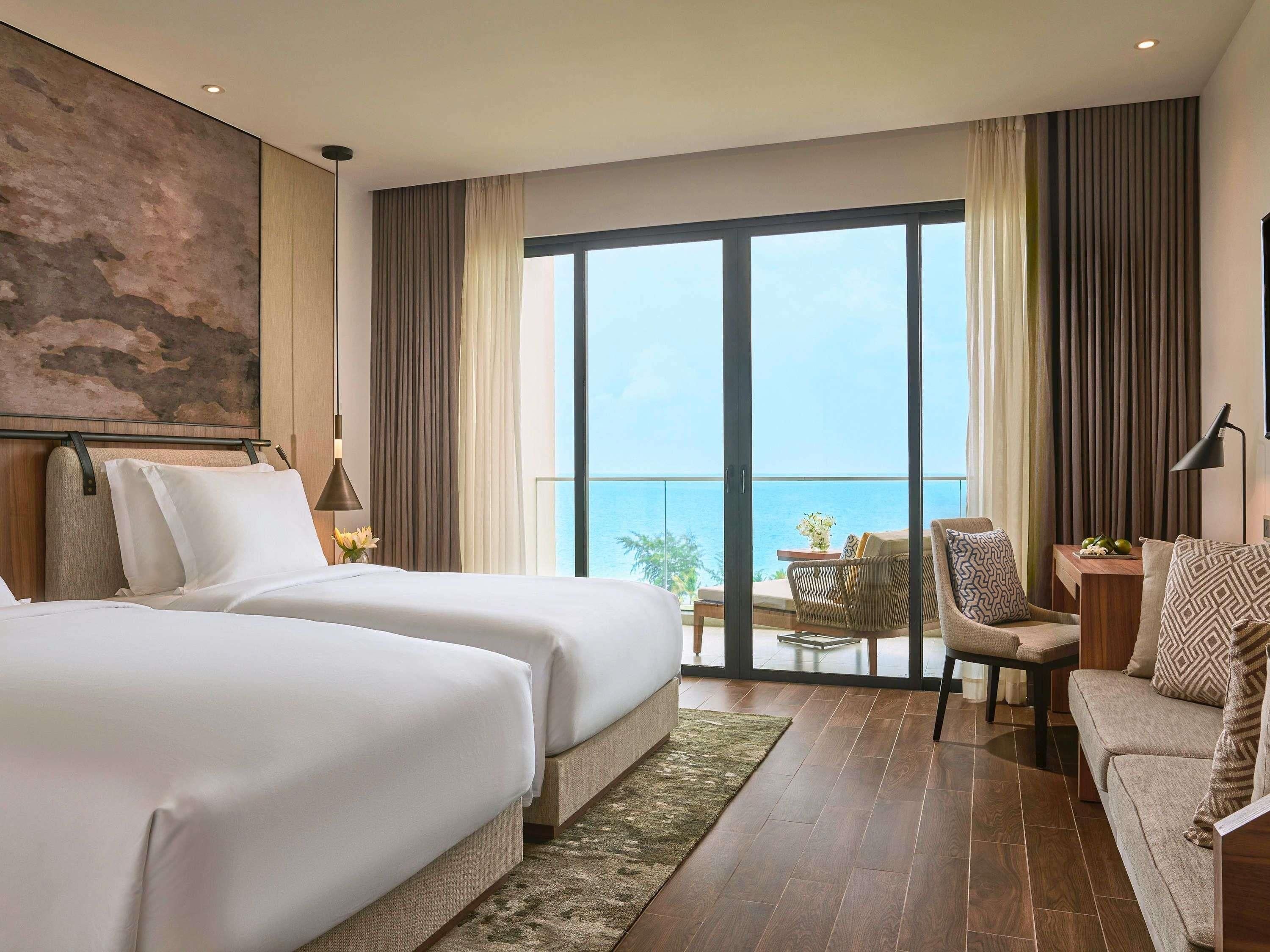 Habitación de hotel con dos camas, un balcón con vistas y una zona de estar con mesa y sillas.