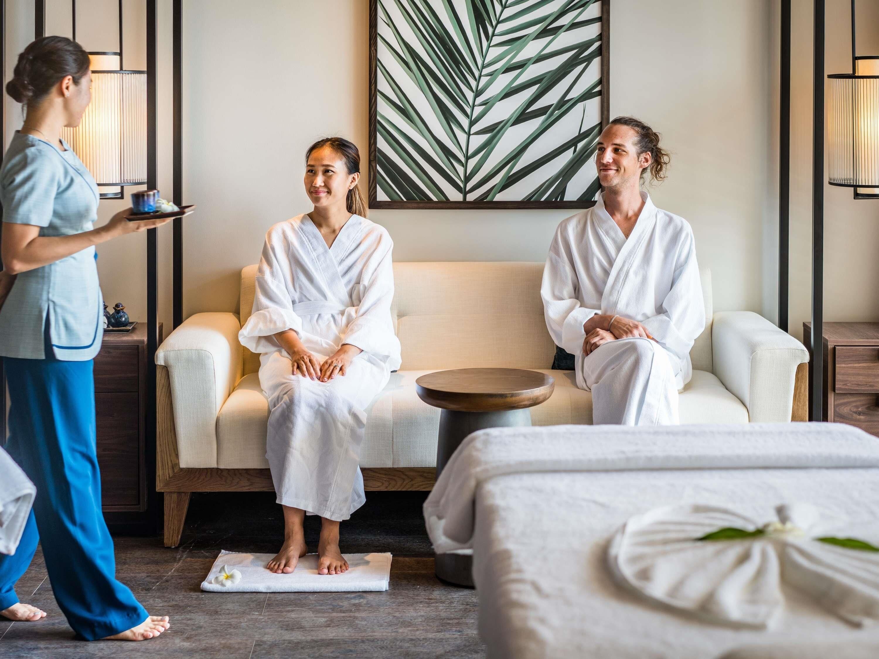 A spa room with two people in white robes, a massage table, a massage table with a flower, and a wall with a leaf pattern.