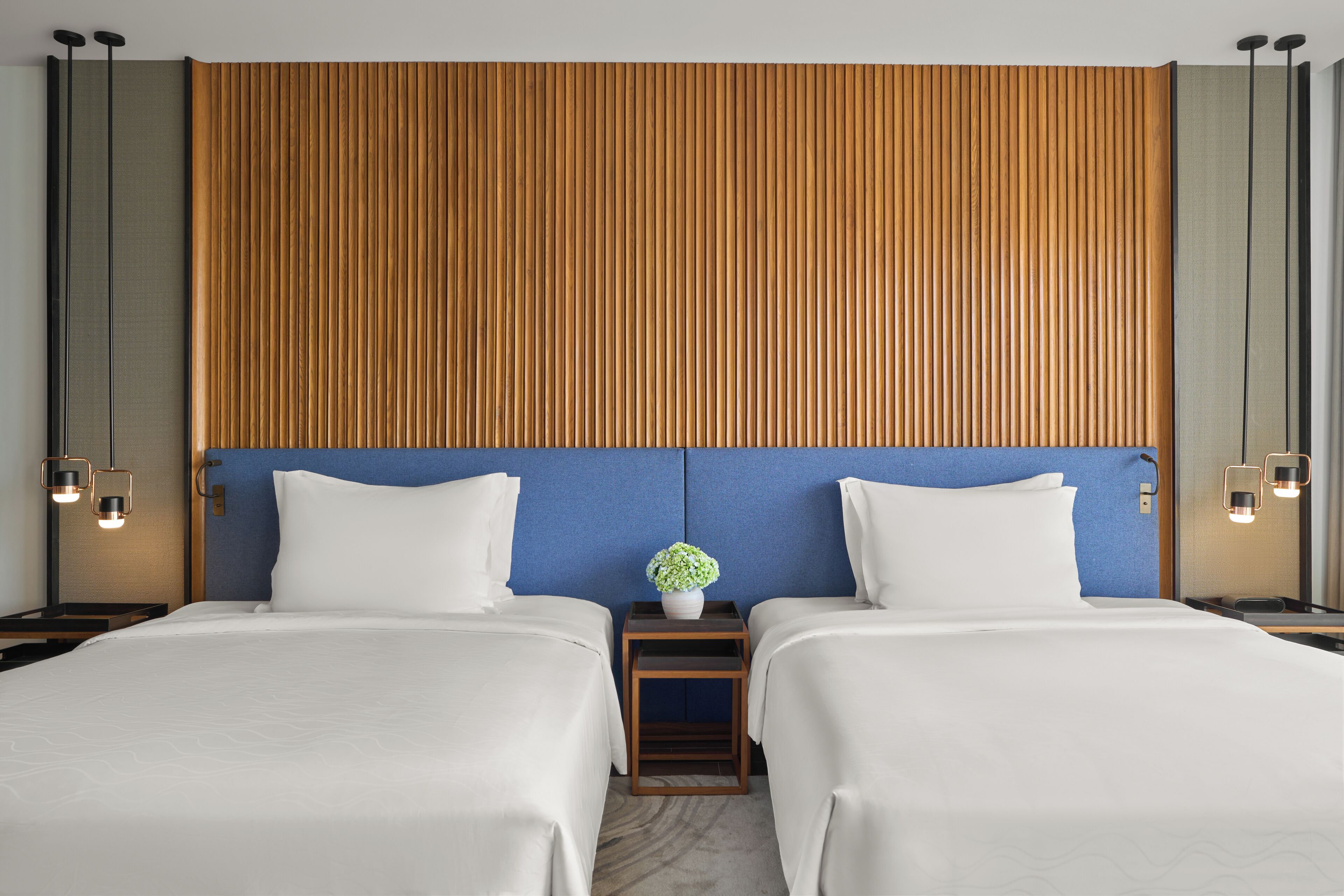 A hotel room with two beds, a wooden headboard, a blue accent wall, and a small table with a vase of flowers.