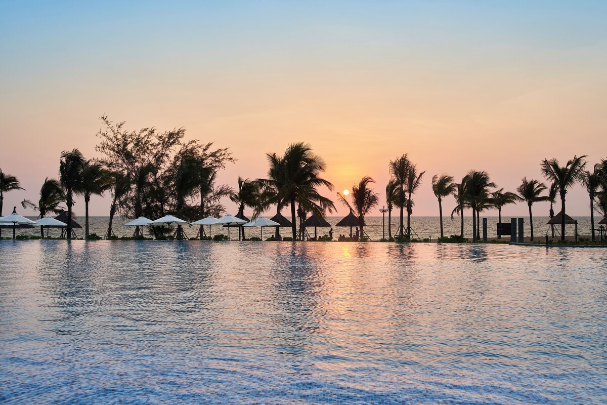 A poolside view at sunset with palm trees and umbrellas.