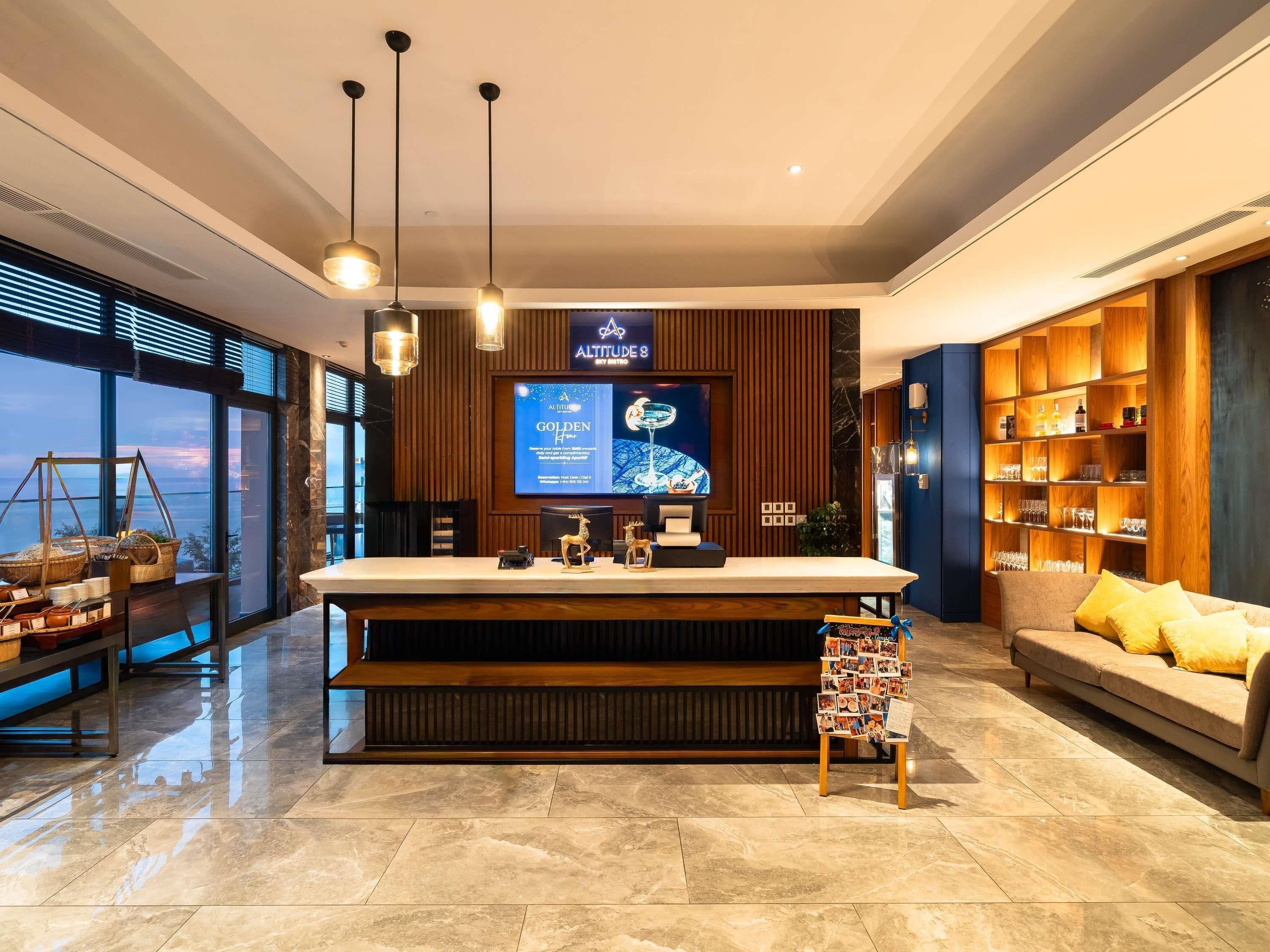 A modern hotel lobby with a reception desk, a large screen displaying the hotel's name, a wooden bookshelf with glasses, and a sofa with yellow cushions.