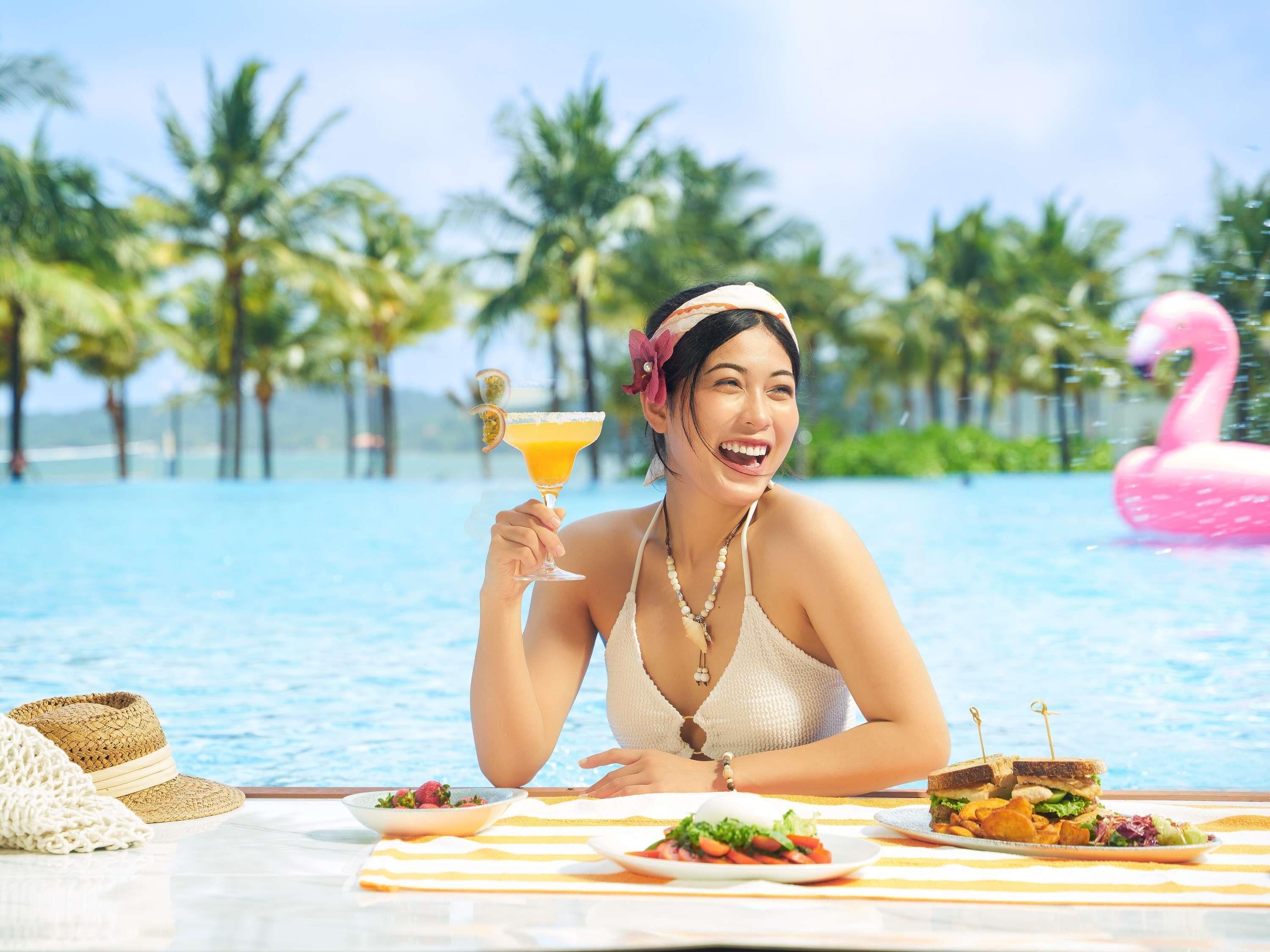 Uma pessoa em uma mesa à beira da piscina, com bebida, comida e um flutuador em formato de flamingo rosa.