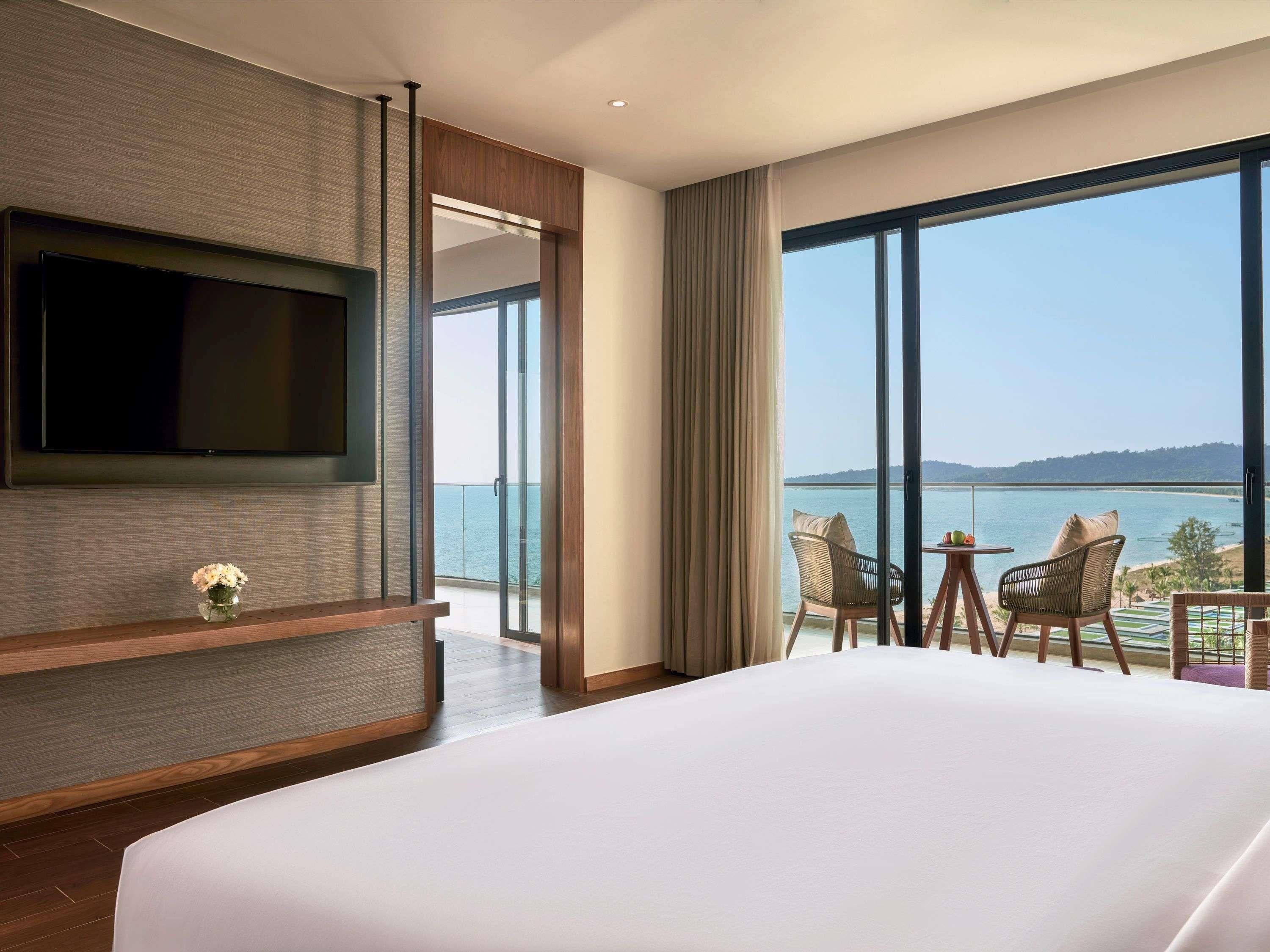 Habitación de hotel con una cama grande, un televisor de pantalla plana en la pared, un balcón con mesa y sillas, y vista al mar.