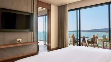 Habitación de hotel con una cama grande, un televisor de pantalla plana en la pared, un balcón con mesa y sillas, y vista al mar.