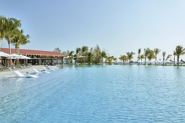 A large swimming pool with lounge chairs and umbrellas, surrounded by palm trees and a building with a red roof. - Mövenpick Resort Waverly Phu Quoc (Phu Quoc)
