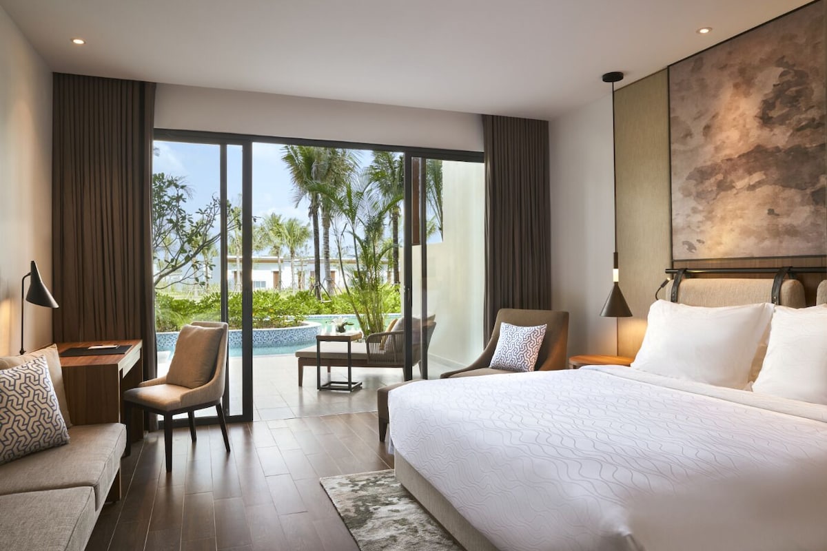 A modern hotel room with a large bed, a desk, a chair, and a view of a pool and palm trees.