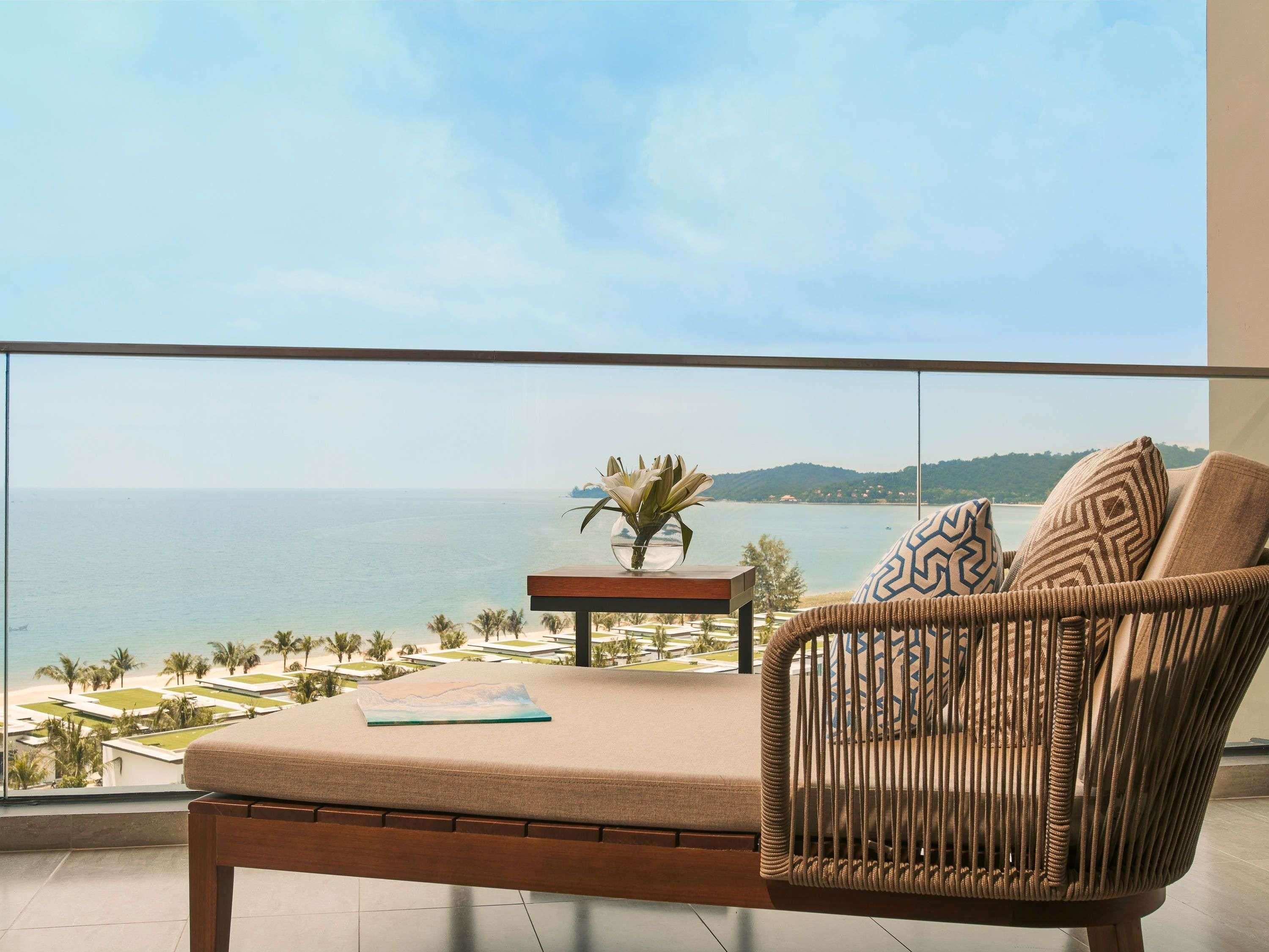 A balcony with a wicker chair, a small table with a plant, and a view of the sea and landscape.