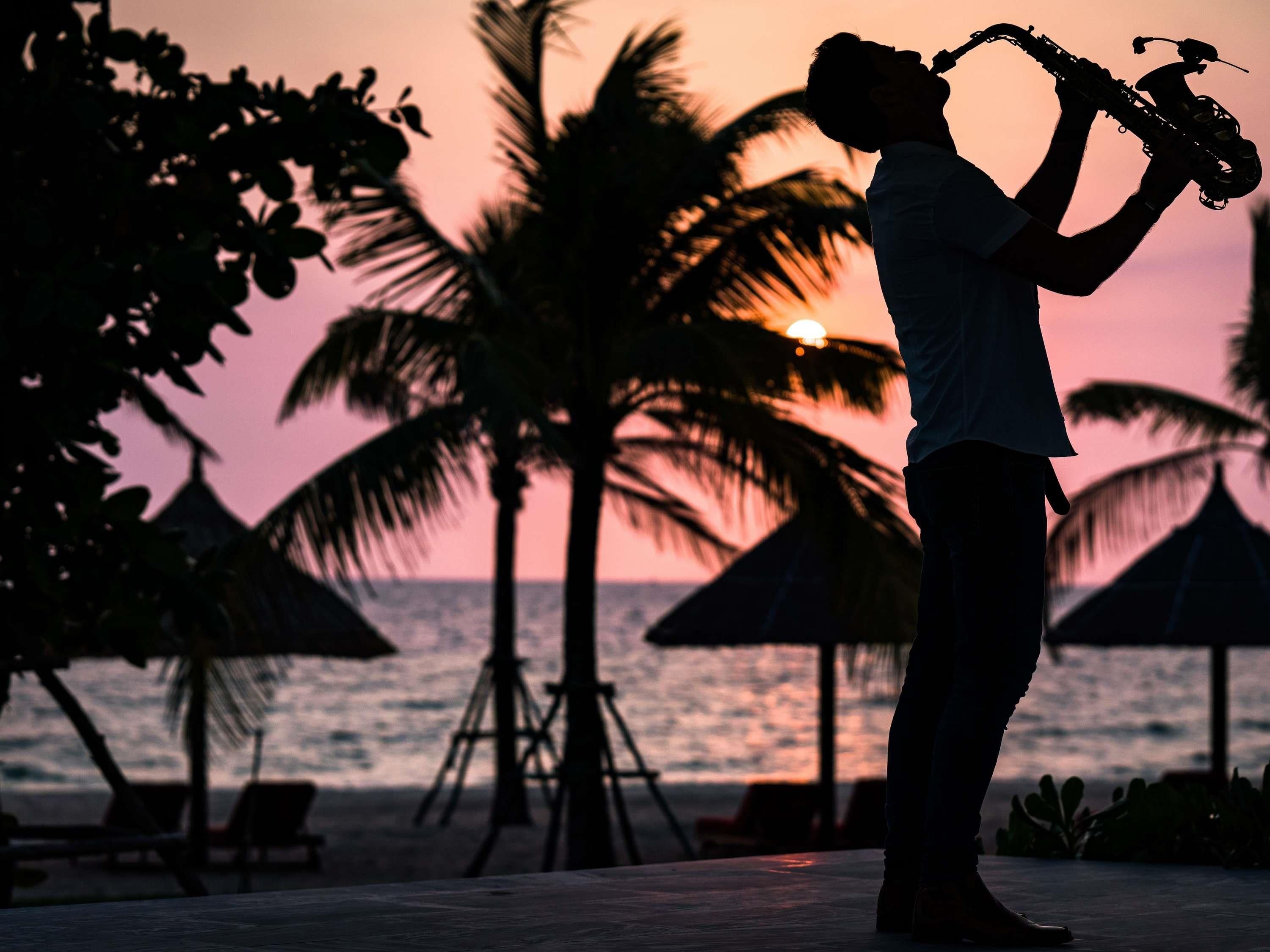 Silueta de una persona tocando un saxofón al atardecer, con palmeras y sombrillas de playa al fondo.