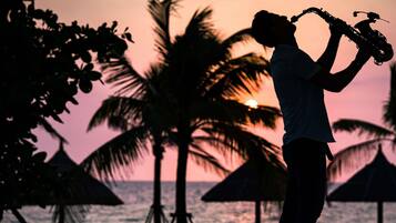 Silueta de una persona tocando un saxofón al atardecer, con palmeras y sombrillas de playa al fondo.