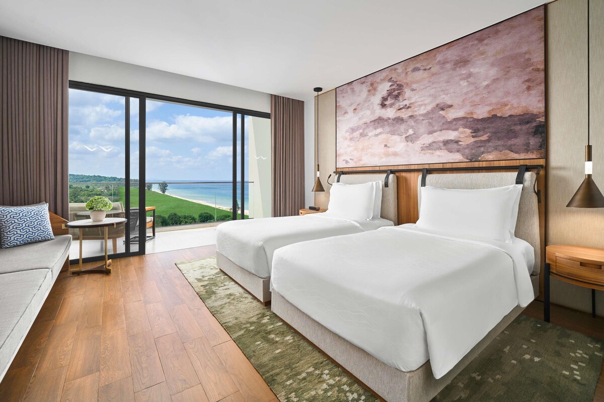 A hotel room with two beds, a sofa, a view of the beach, and a large abstract painting on the wall.