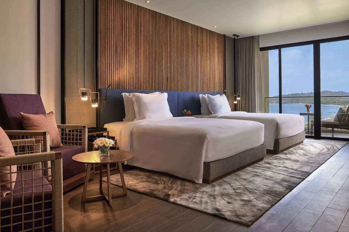 A hotel room with two beds, a view of the sea, a balcony, and a dining area with a table and chairs.