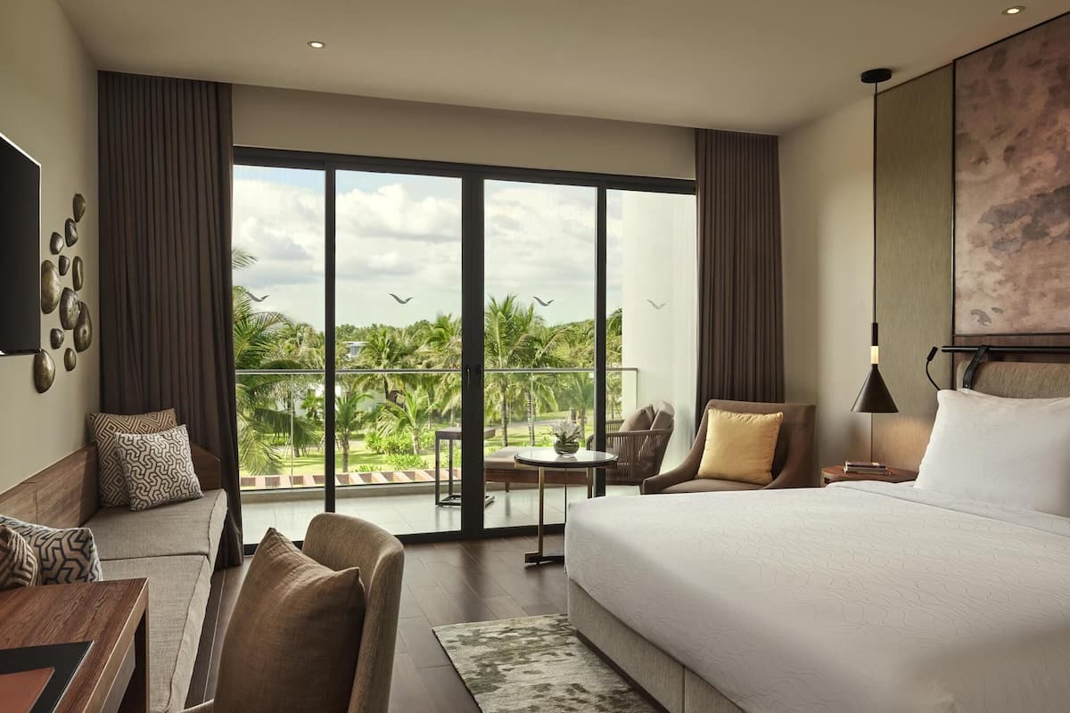 A hotel room with a large bed, a sofa, a desk, a chair, and a balcony with a view of palm trees and the sky.