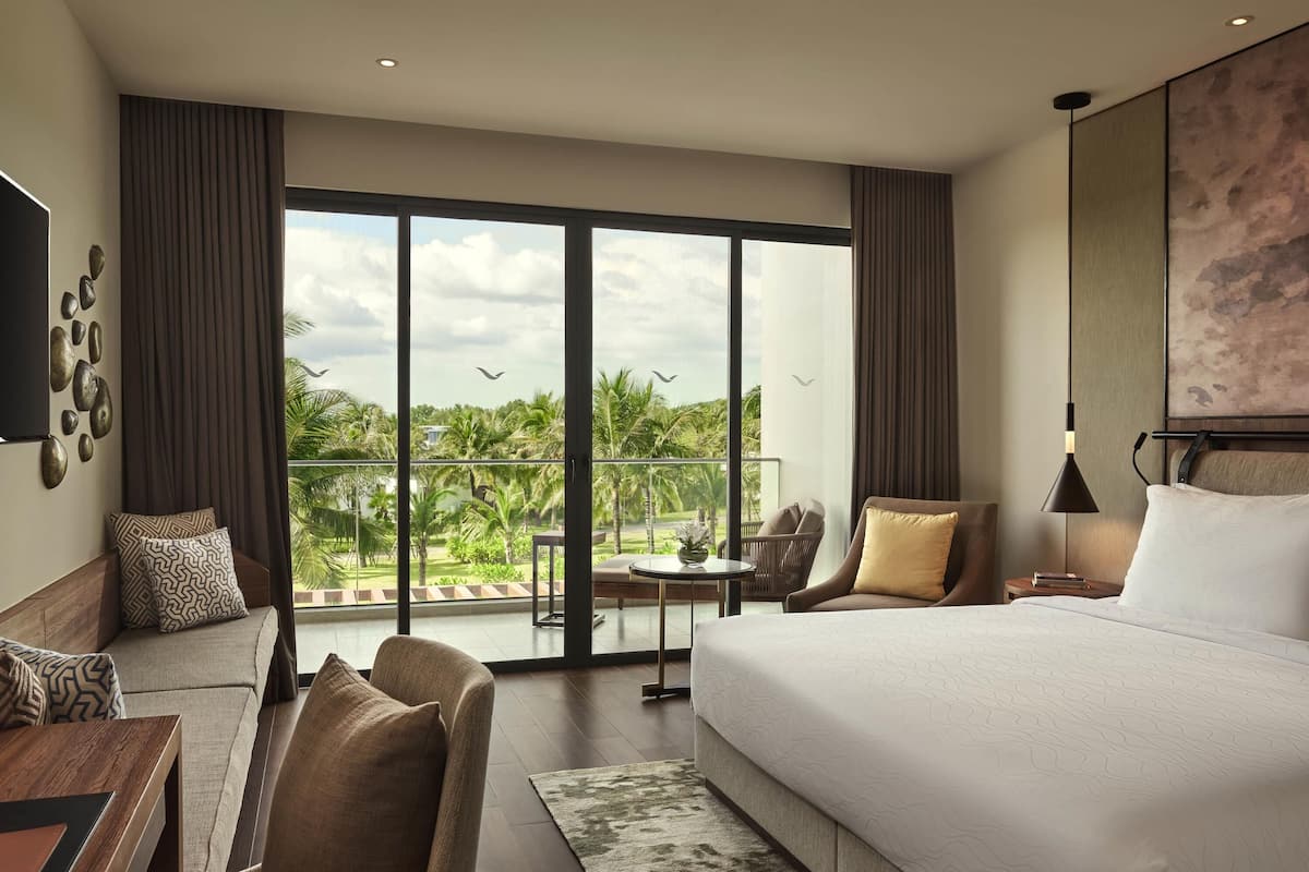 A hotel room with a large bed, a sofa, a desk, and a balcony with a view of palm trees and the sky.