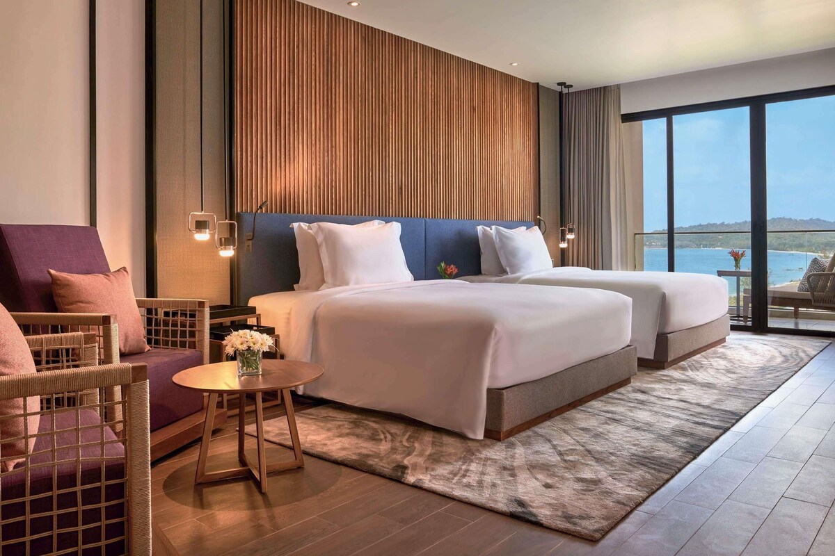 A hotel room with a large bed, a sofa, a small table, and a view of the sea.