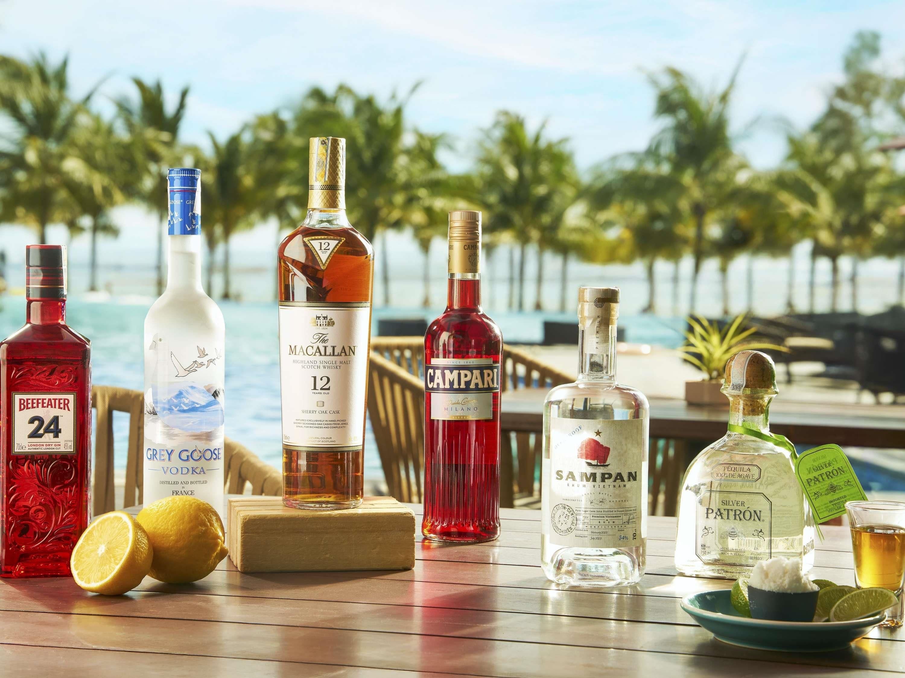 A variety of spirits and a glass of beer on a wooden table by a poolside with palm trees in the background.