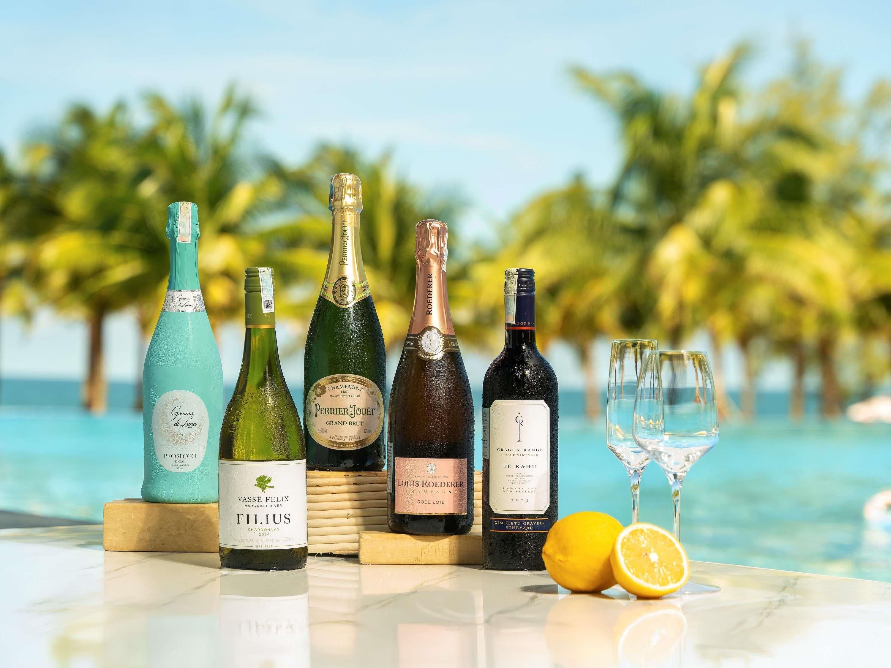 A selection of wine bottles and glasses on a table with a pool and palm trees in the background.