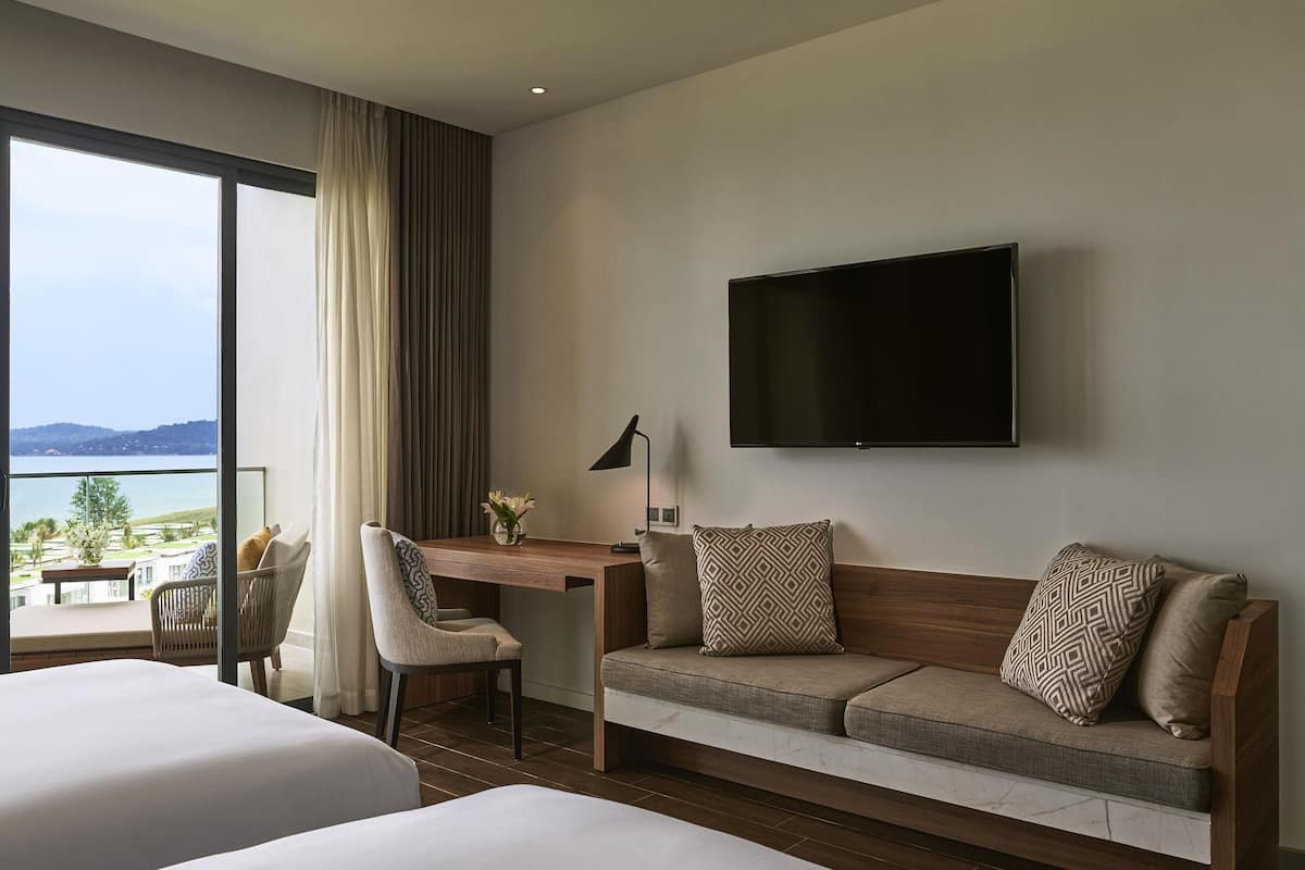 A hotel room with two beds, a sofa, a desk, a chair, a TV, and a view of the sea.