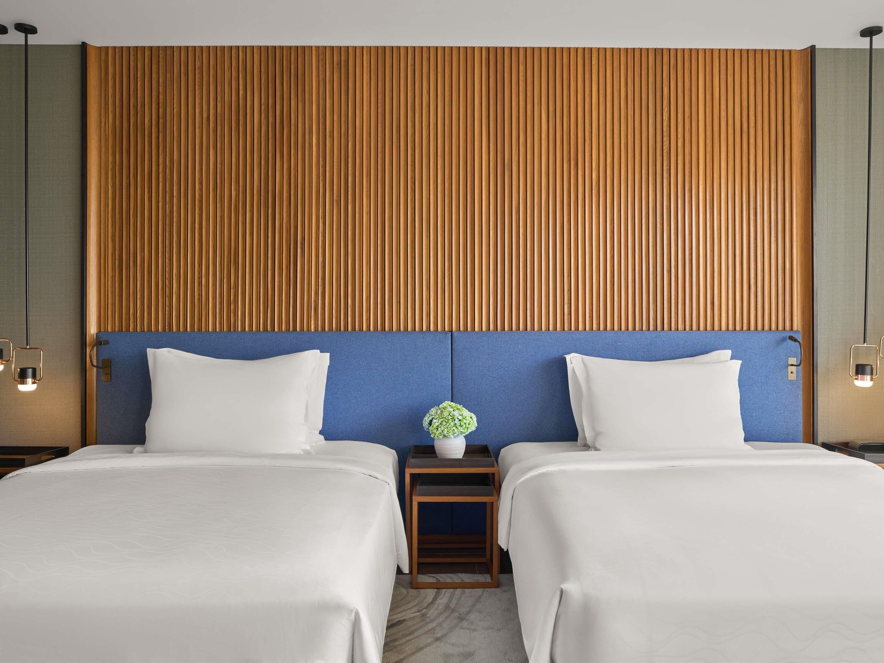 A hotel room with two beds, a wooden slatted headboard, a small bedside table with a plant, and wall-mounted lights.