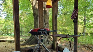 Un juego de parrilla portátil Coleman instalado en un área boscosa, con una estructura de madera encima, un carrito de parrilla metálico y un farol colgante.