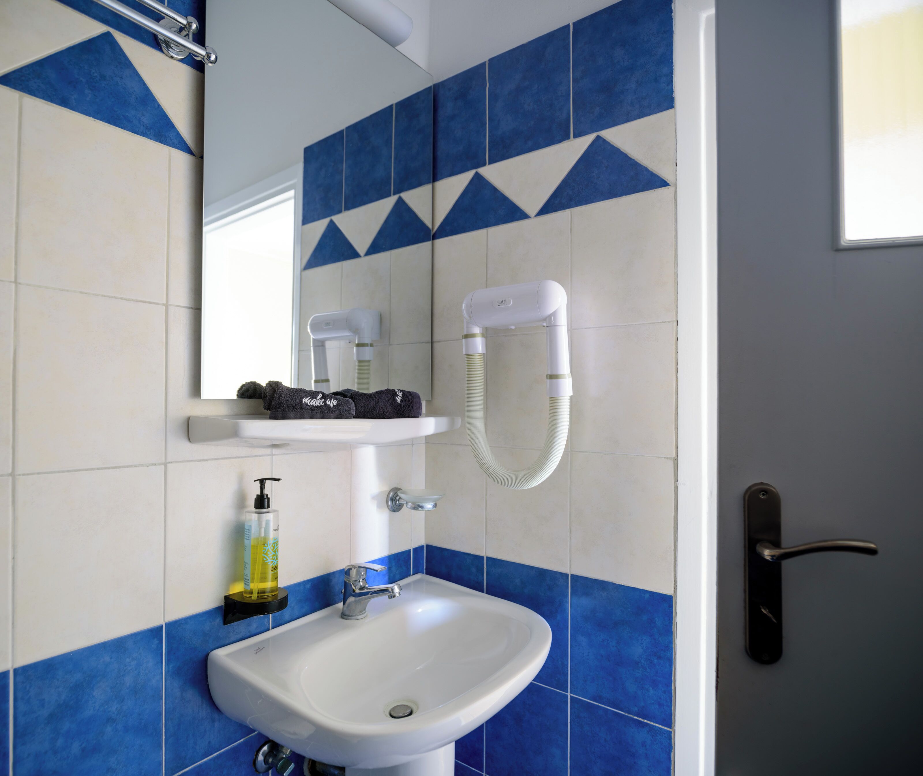 Un baño con lavamanos blanco, azulejos azules y blancos, un perchero, un secador de pelo y una botella de jabón para manos.