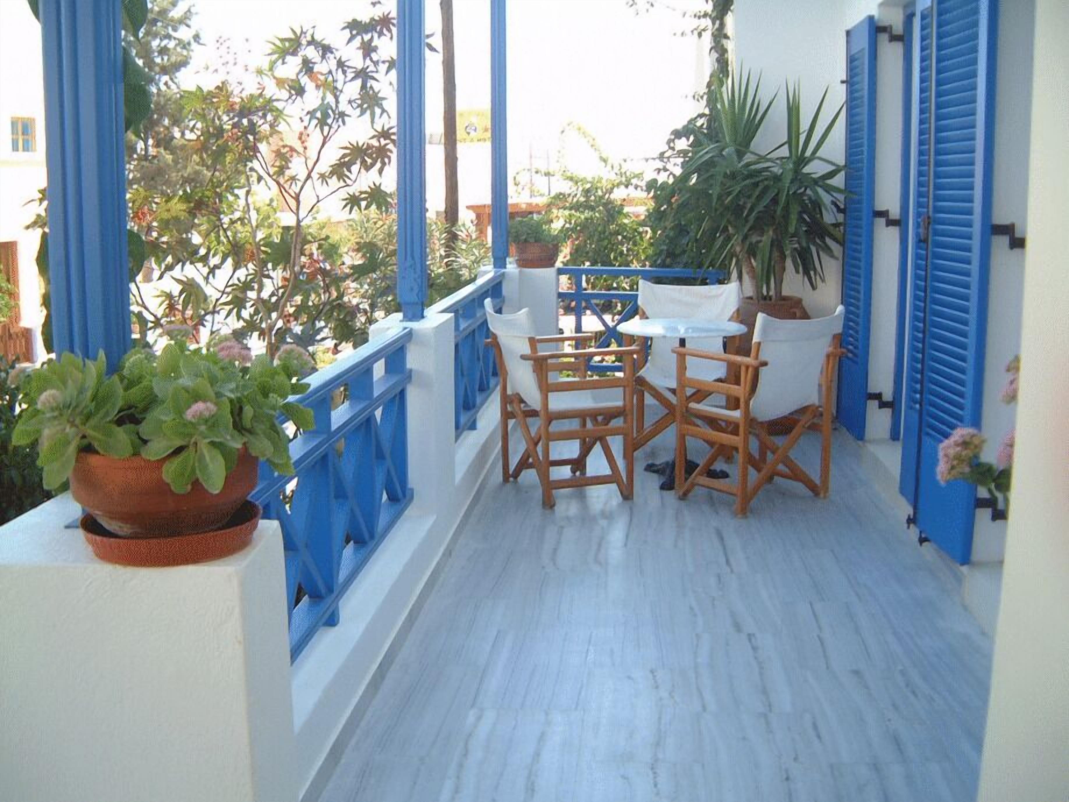 A balcony with blue railings, white walls, and a table with chairs. There are potted plants and a large tree in the background.