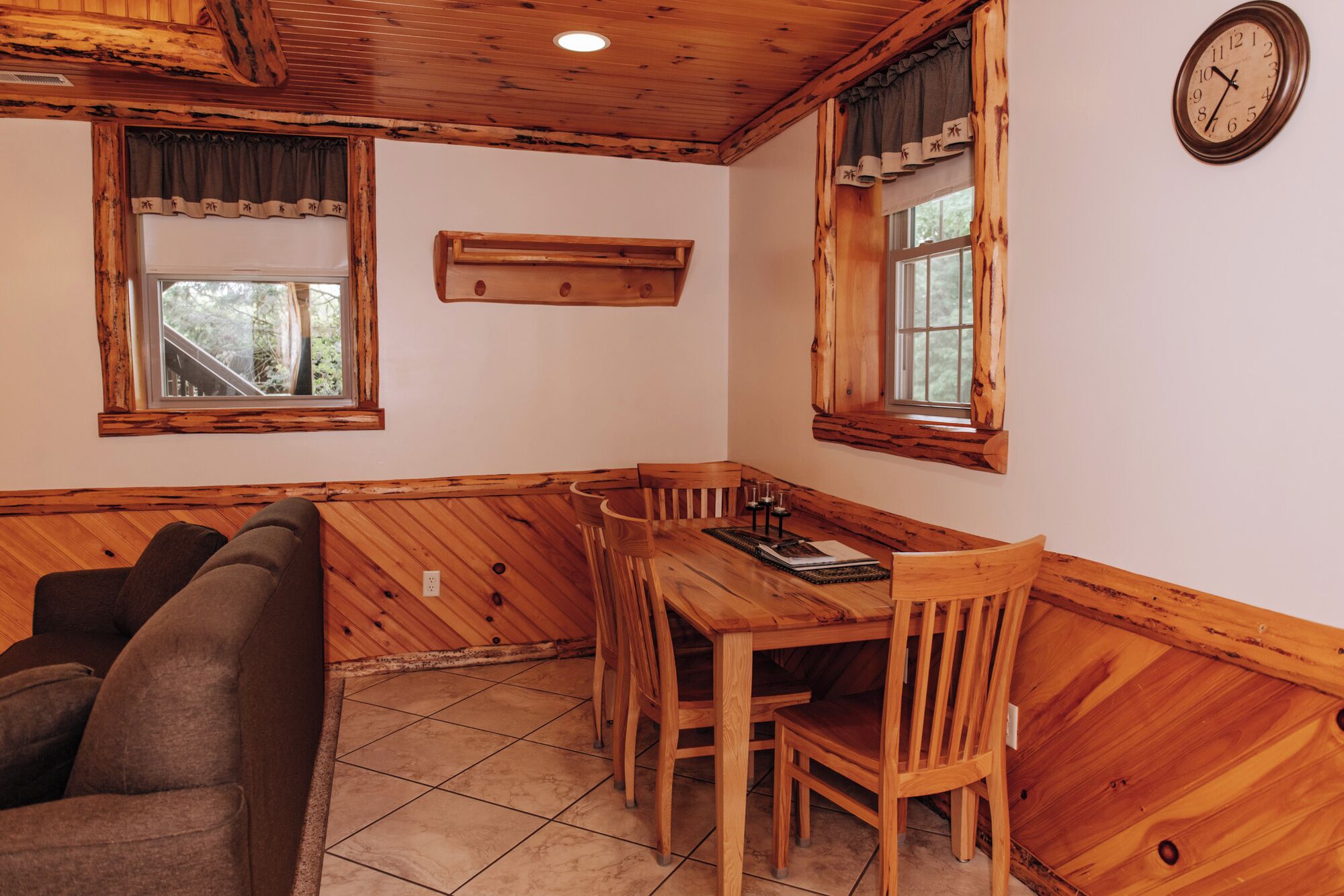 A cozy room with a wooden ceiling, a dining table with chairs, a sofa, and two windows with curtains.