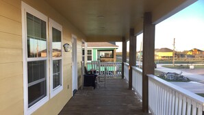Une véranda couverte avec une terrasse en bois, une balustrade et une vue sur une maison verdoyante en arrière-plan.