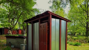 A small, single-story shed with a red door and green siding, located in a grassy area with trees in the background.