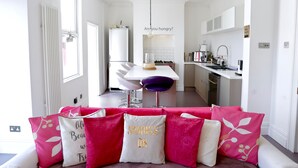 A modern kitchen with white cabinets, a dining area with a white table and purple chairs, and a sofa with pink and white cushions. - Harrington House (Bristol)