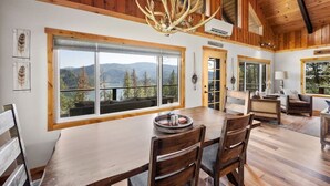 A spacious dining area with a large wooden table and chairs, a chandelier, and large windows offering a view of mountains and trees.