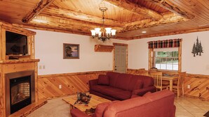 Interior acolhedor de uma cabana com lareira, paredes de madeira, um sofá, uma mesa de jantar e uma janela com vista para árvores.