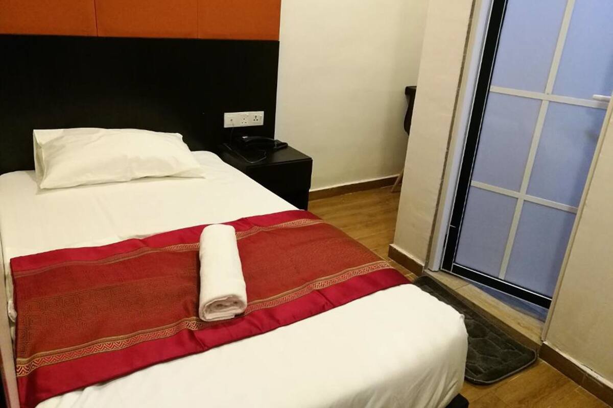 Cama bem arrumada com colcha vermelha e branca, um travesseiro branco e uma toalha branca enrolada. Tem uma mesa de cabeceira preta com um telefone e uma tomada. Uma janela mostra vista do céu azul e de um edifício.