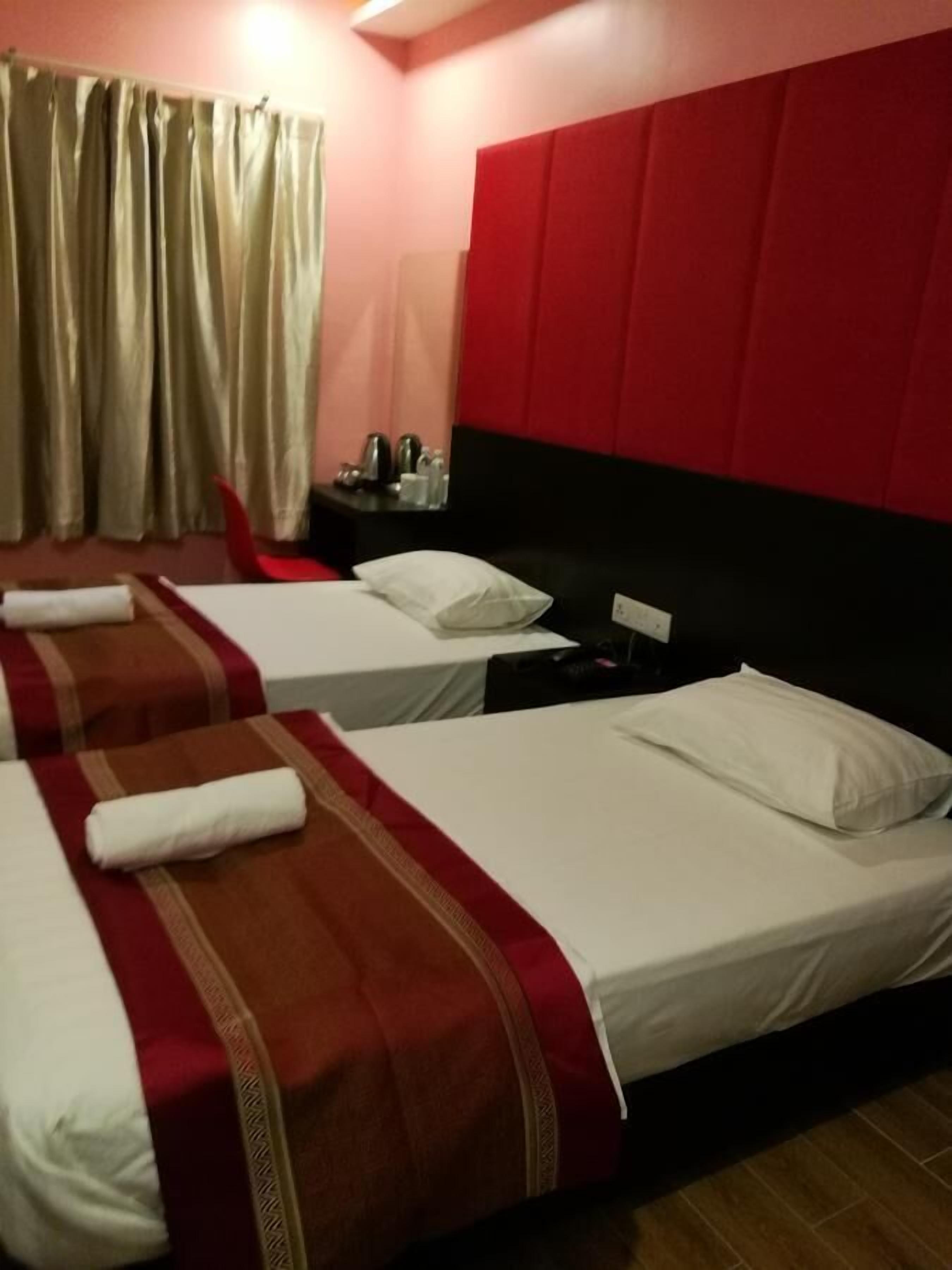 A hotel room with three beds, each with white linens and red and brown decorative pillows. There is a nightstand with a lamp and a small table with a few items on it. The room has a red headboard and beige curtains.