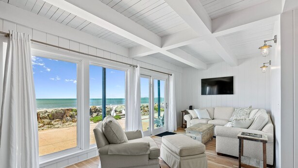 Amplia sala de estar con grandes ventanales que dan a una playa, un sofá color beige y piso de madera.