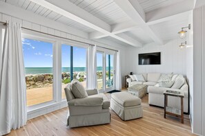 A spacious living room with large windows overlooking a beach, a beige sofa, and a wooden floor. - Casa Del Mar in Rio del Mar! (Aptos)