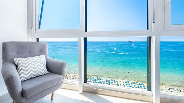 Una stanza con una grande finestra vista spiaggia, una poltrona grigia con cuscino decorato e un pavimento piastrellato.