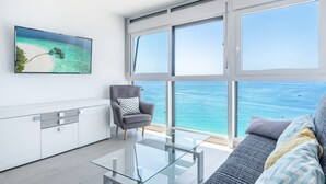 Un soggiorno moderno con una grande finestra vista mare, una TV a schermo piatto a parete, una poltrona grigia e un tavolino da caffè in vetro.