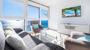 Una sala de estar moderna con un amplio ventanal con vista al mar, un sofá confortable, una mesa de centro de vidrio y un mueble blanco minimalista.