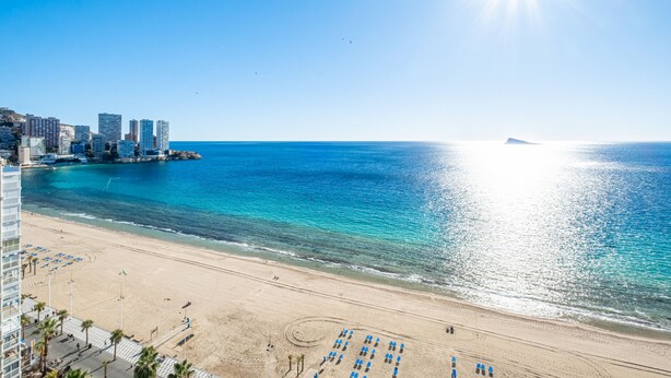 Una playa con un paisaje urbano al fondo, cielo despejado y sol brillante.