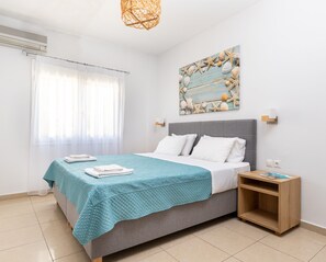 A bedroom with a bed, a nightstand, a wall-mounted light, a ceiling fan, and a beach-themed artwork. - Sun Beach Apartments (Chania)