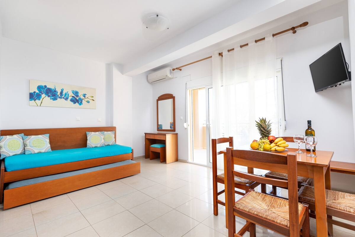 A modern room with a wooden sofa, a dining table with chairs, a fruit bowl, and a flat-screen TV.
