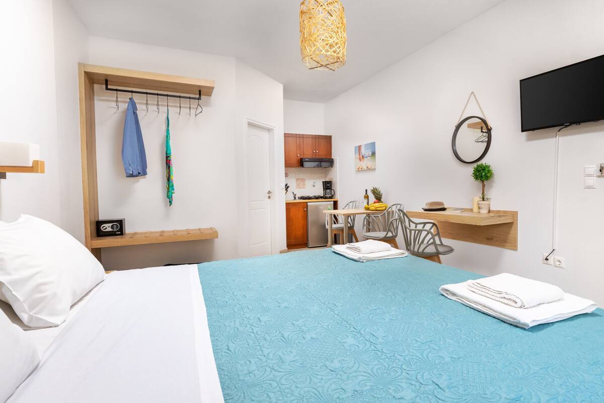 A hotel room with a bed, a desk with a chair, a small kitchenette, and a TV on the wall.
