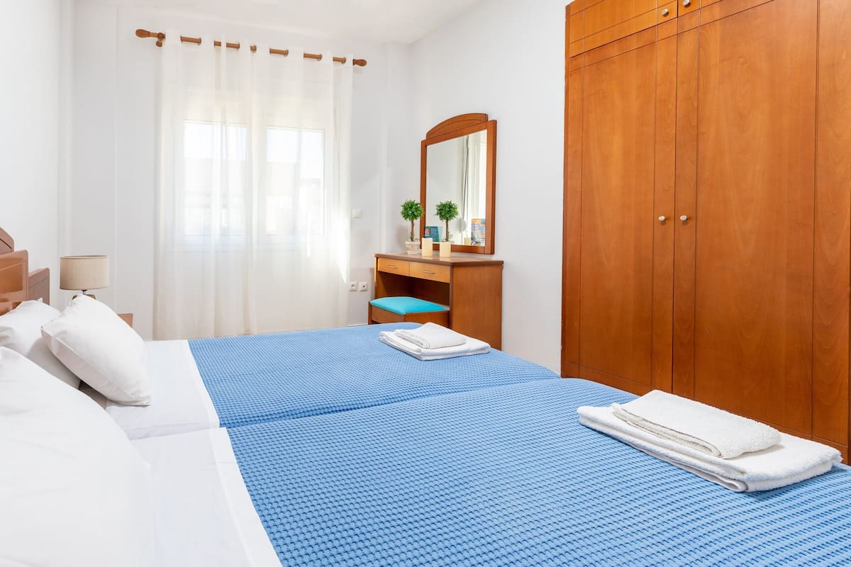 A hotel room with a bed, a wooden wardrobe, a desk with a mirror, and a window with sheer curtains.