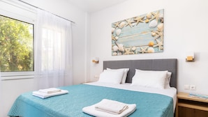 A neatly made bed with a teal bedspread and white pillows, a wall-mounted artwork of seashells and starfish, and a window with sheer curtains. - Sun Beach Apartments (Chania)