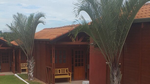 A row of wooden buildings with red-tiled roofs, each with a bench and a palm tree in front. - Pousada Ilson (Olímpia)