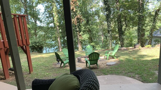 A covered patio area with wicker furniture and green chairs, overlooking a grassy yard with trees and a body of water. - Quiet sunsets on Lanier (Oakwood)