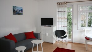 A living room with a grey sofa, red cushions, a white TV stand, a black chair, a small white table, and a red rug. - Guest house Villa Lina (Bad Herrenalb)