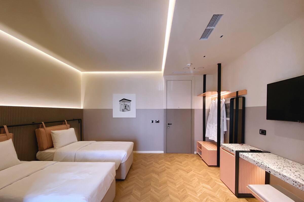 Une chambre d’hôtel avec deux lits, un parquet en bois disposé en point de Hongrie, une salle de bain avec un miroir et un téléviseur fixé au mur.