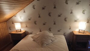 A bedroom with a sloped ceiling, a bed with white bedding, two bedside tables with lamps, and a wall with a bamboo plant pattern. - Chalet 6/7 people at the foot of the Pyrenees 2000 slopes in the forest (Bolquère)