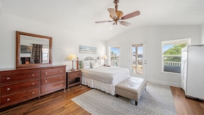 A bedroom with a bed, dresser, bedside lamps, a ceiling fan, a bench, a large window, and a door leading to a balcony.
